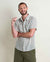 Man wearing light blue plaid short-sleeve button-up shirt with green pants, smiling