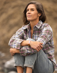 Woman seated outdoors wearing a plaid flannel button-up shirt with rolled jeans, looking thoughtfully upward