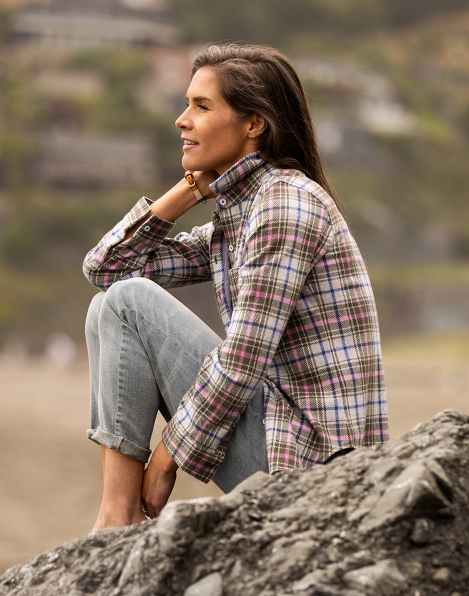 Woman wearing a plaid flannel button-up shirt with rolled jeans, seated on rocks outdoors