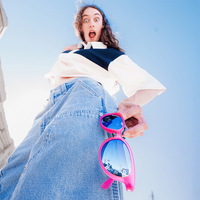 Person holding oversized pink-framed sunglasses with blue mirrored lenses toward camera from below