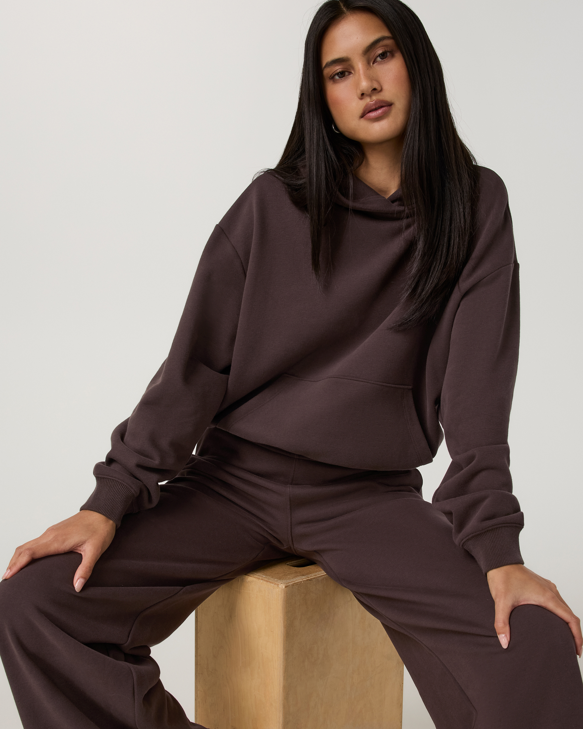 Women's pullover hoodie in dark brown, seated model showing front view and matching joggers
