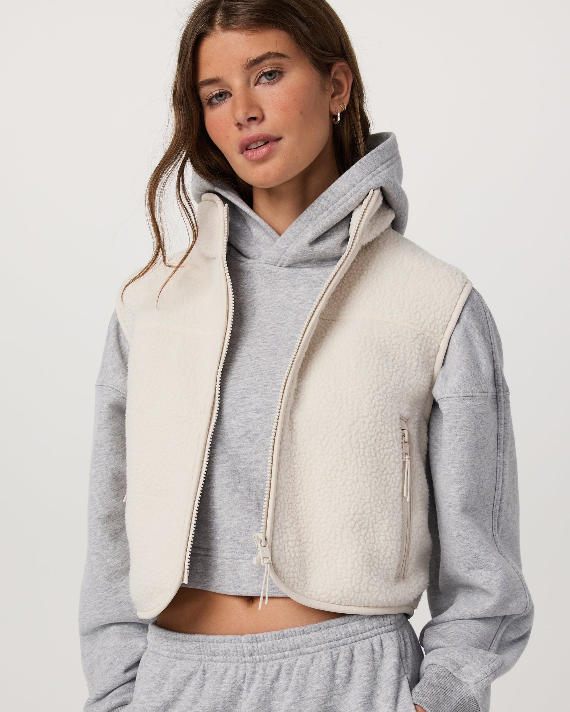 Women's cropped sherpa fleece vest with asymmetric zip, worn over gray hoodie, front view