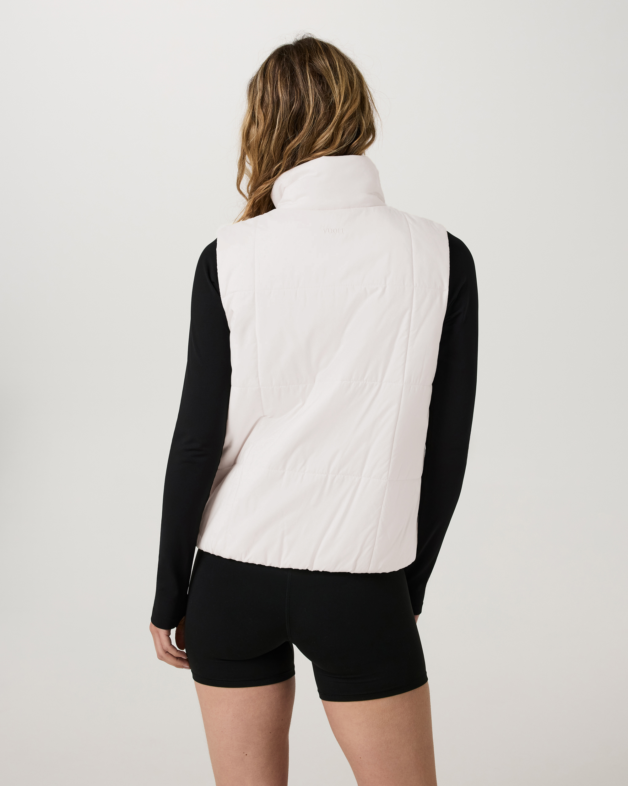 Women's insulated puffer vest in pale pink, back view, worn over black long-sleeve top