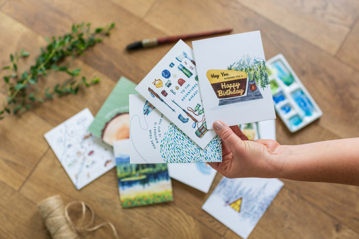 Hand holding greeting cards with watercolor designs including birthday and adventure themes on wooden surface