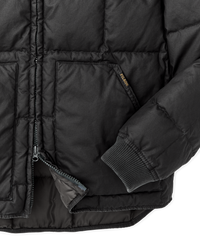 Close-up detail of quilted black down jacket showing zipper, pocket, ribbed cuff, and brand label