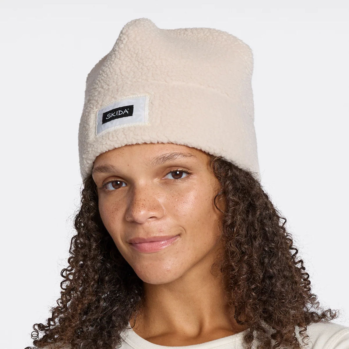 Woman wearing a cream high-pile sherpa beanie with brand label patch, front view
