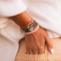 Stacked hair tie bracelets on wrist featuring orange, blue, and blush braided cords with gold wave charm