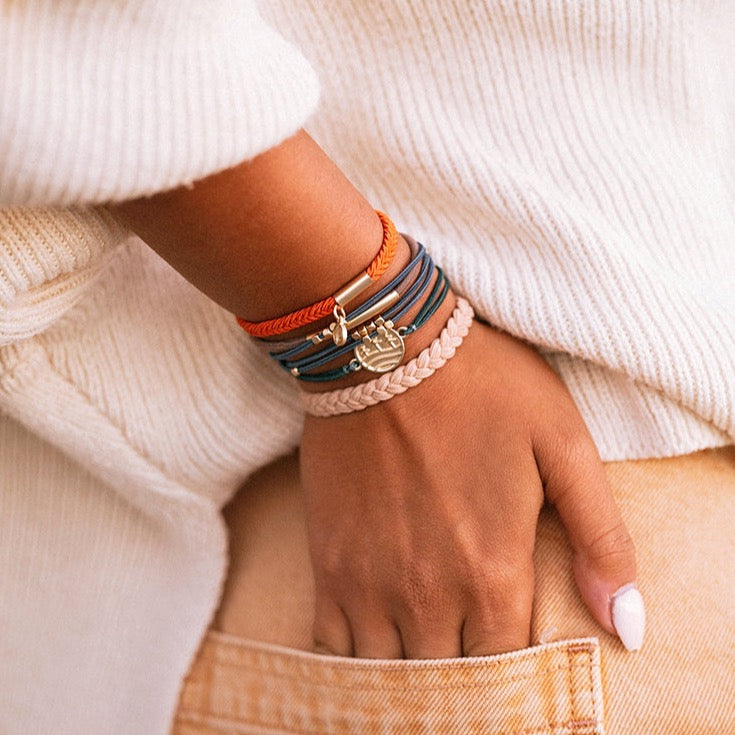 Stacked hair tie bracelets on wrist featuring orange, blue, and blush braided cords with gold wave charm