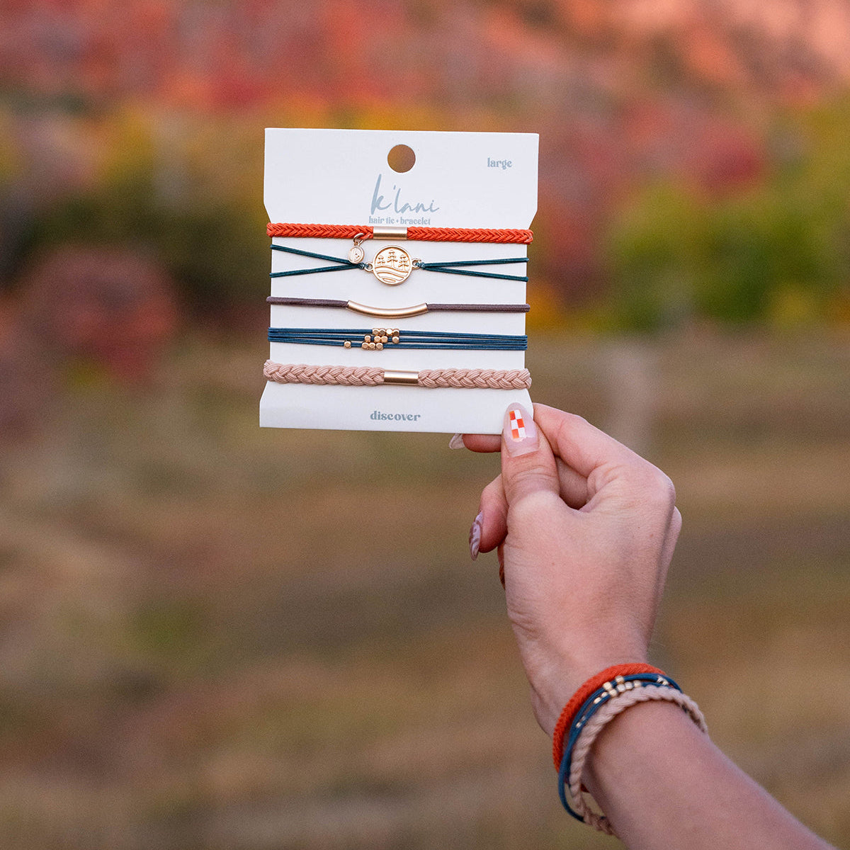Hand holding a card of five stacked hair tie bracelets in earth tones with gold charm accents