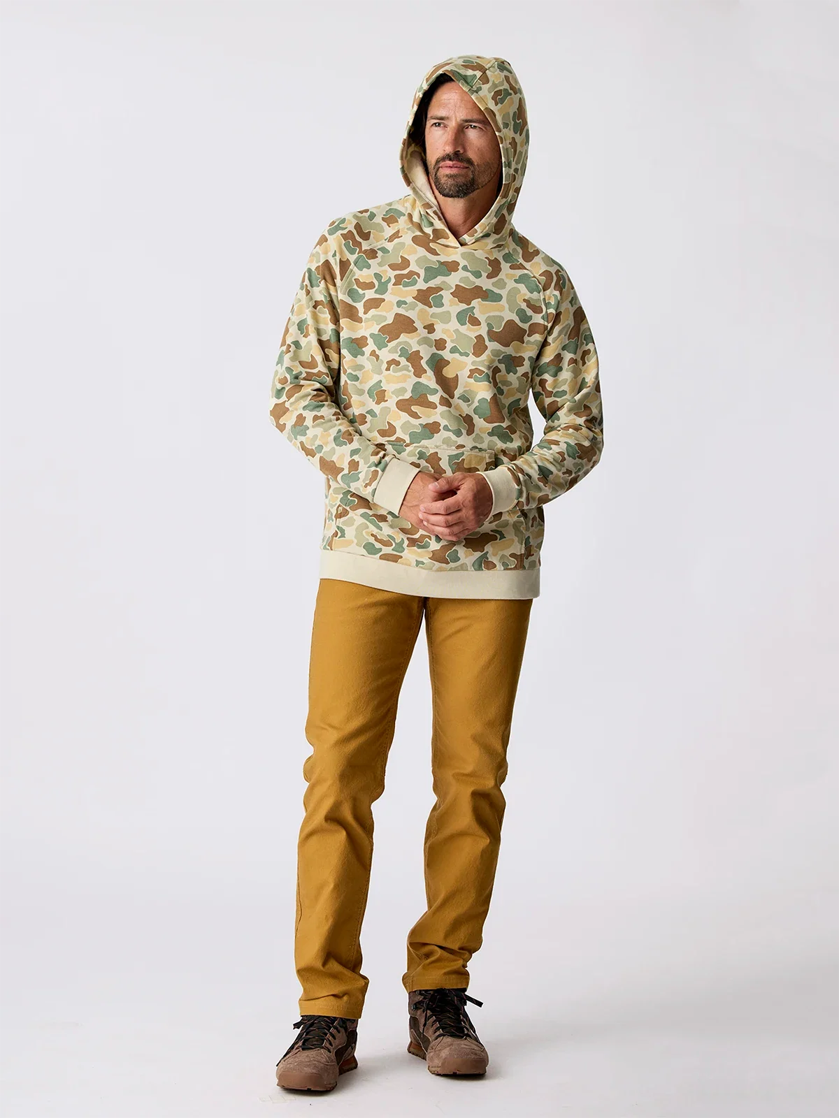 Men's camo pullover hoodie with kangaroo pocket, worn with mustard pants, full body front view