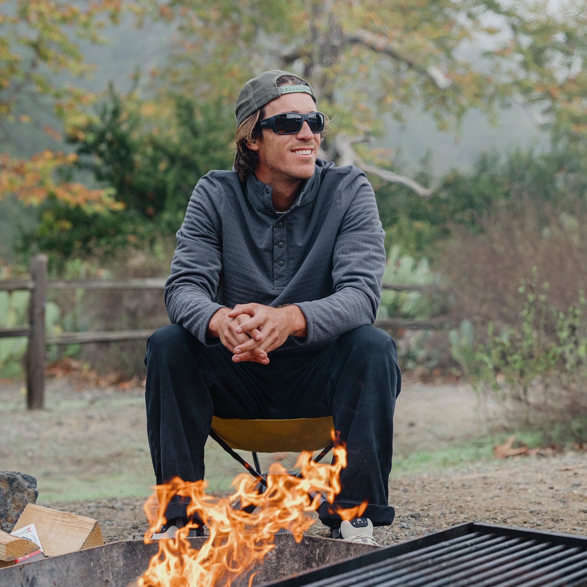 Man wearing black sunglasses sitting by campfire in outdoor setting