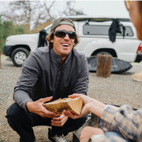 Man wearing black sunglasses outdoors while sharing food from wooden bowl