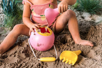 Pink bucket-style beach toy with yellow handle and spade being used in sand by child