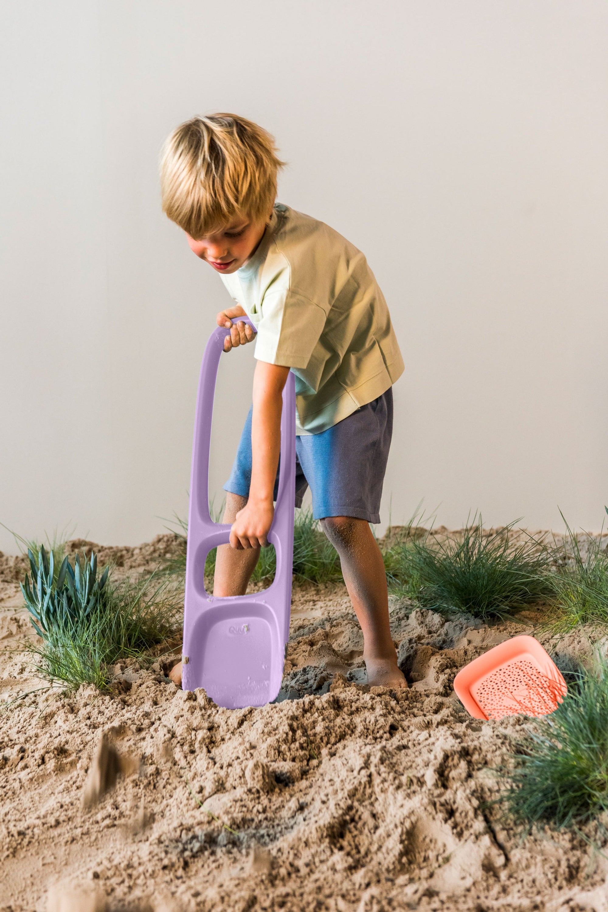 Child using purple sand digger toy in sandy play area with grass patches