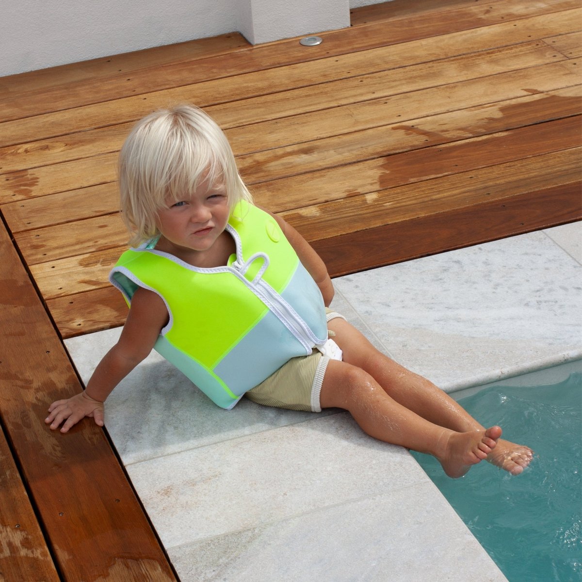 Child wearing bright green and blue swim vest sitting by pool edge with feet in water