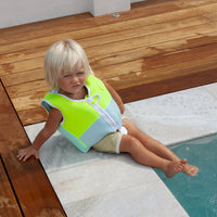 Child wearing bright green and blue swim vest sitting by pool edge with feet in water