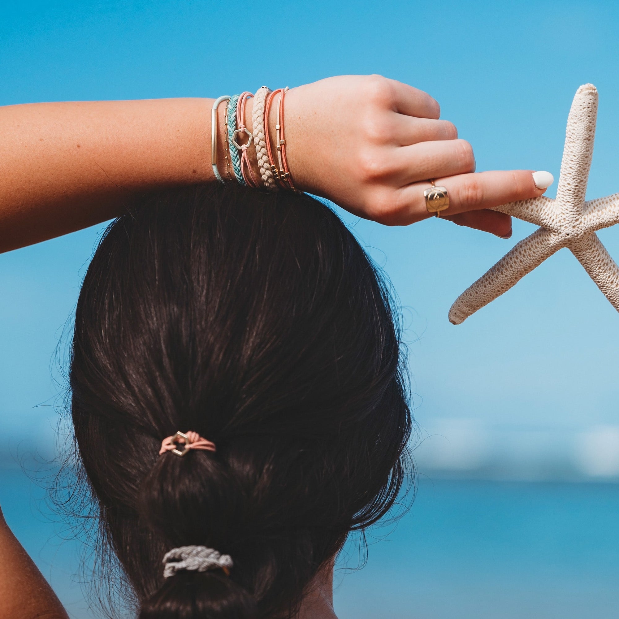 Braided hair tie worn in ponytail with matching bracelet stack and starfish held at beach