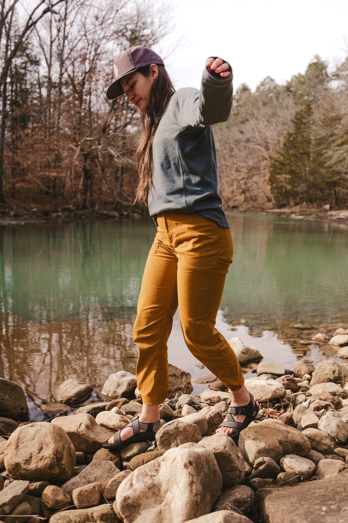 Women's trail pants in warm amber tone, worn on rocky riverbank with sandals, active outdoor setting