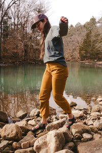 Women's trail pants in warm amber tone, worn on rocky riverbank with sandals, active outdoor setting