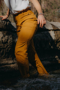 Women's trail pants in golden caramel, side view, worn outdoors near rocky stream