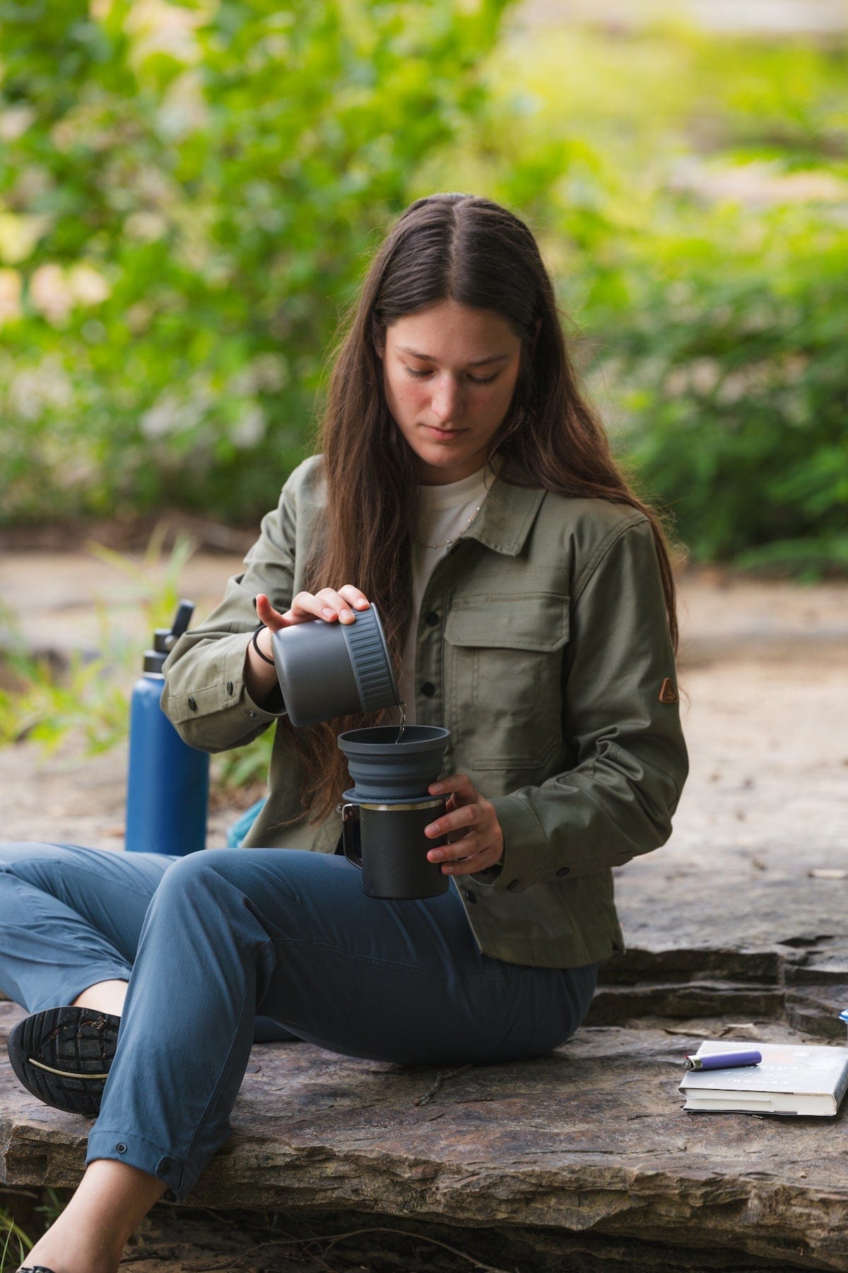 Woman sitting outdoors on rocks wearing an olive button-up shirt jacket with chest pockets