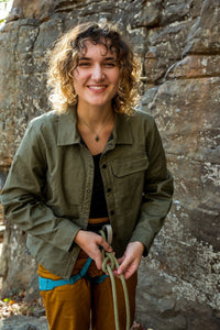 Woman wearing olive green button-up shirt jacket outdoors, holding climbing rope near rock face