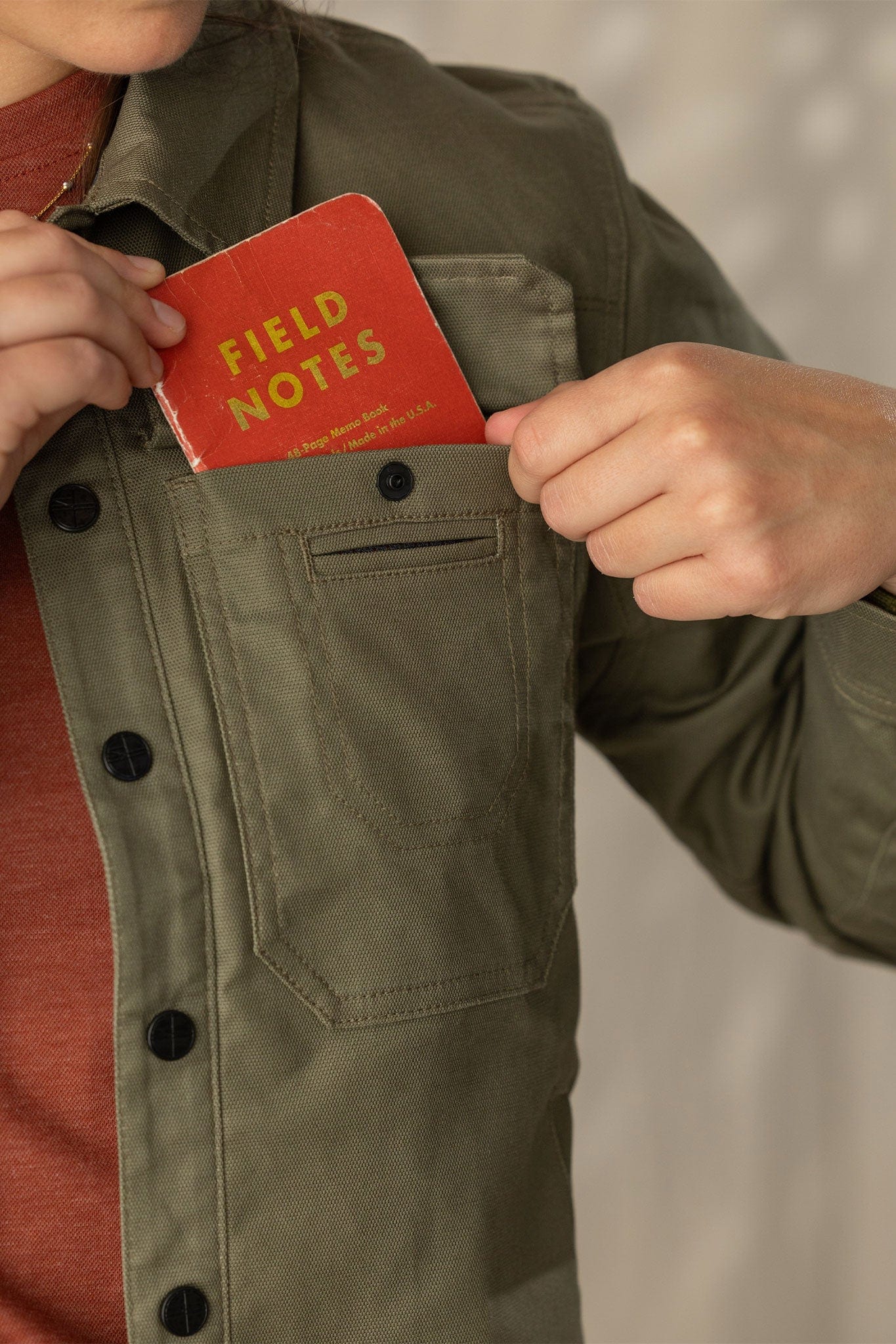 Close-up detail of olive jacket chest pocket with red Field Notes notebook being inserted