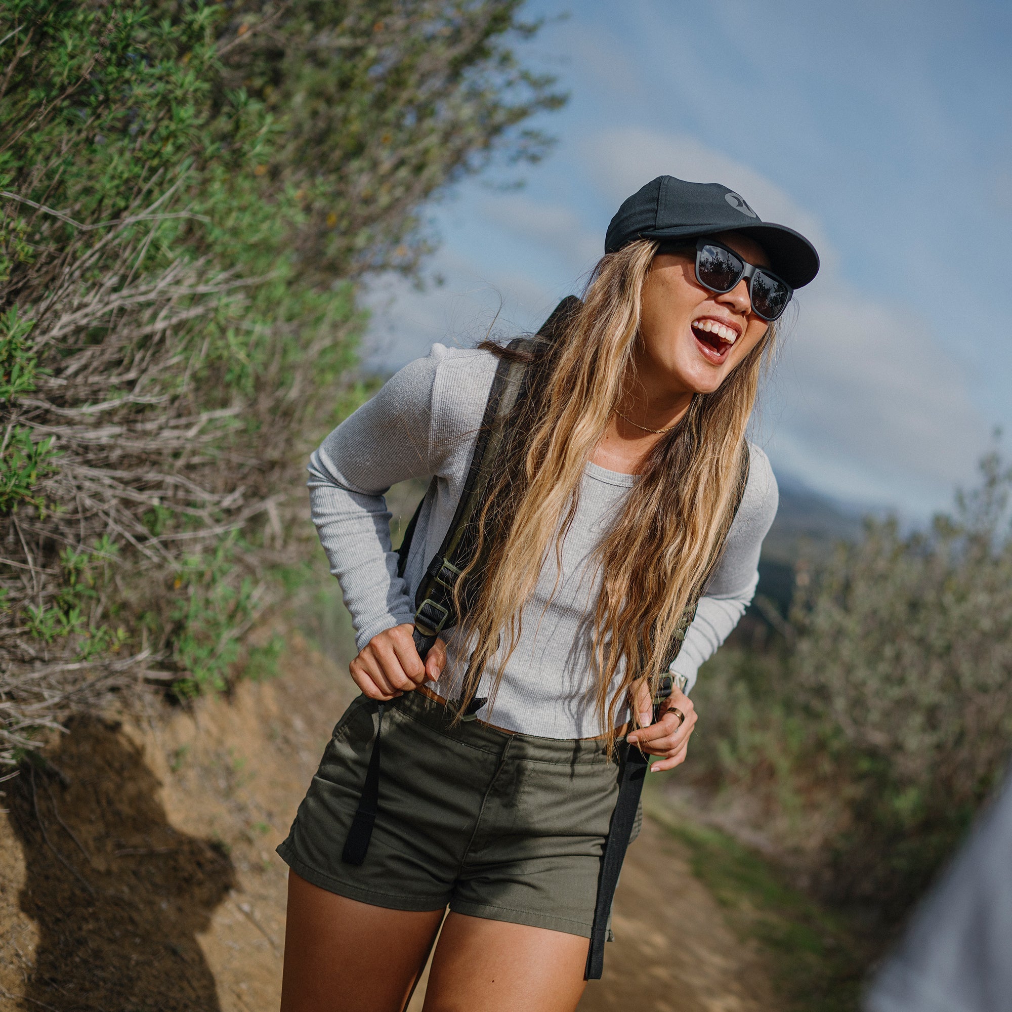 Woman wearing black sunglasses and cap laughing outdoors with backpack straps