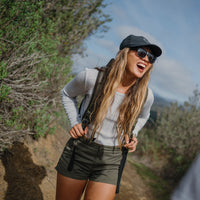Woman wearing black sunglasses and cap laughing outdoors with backpack straps