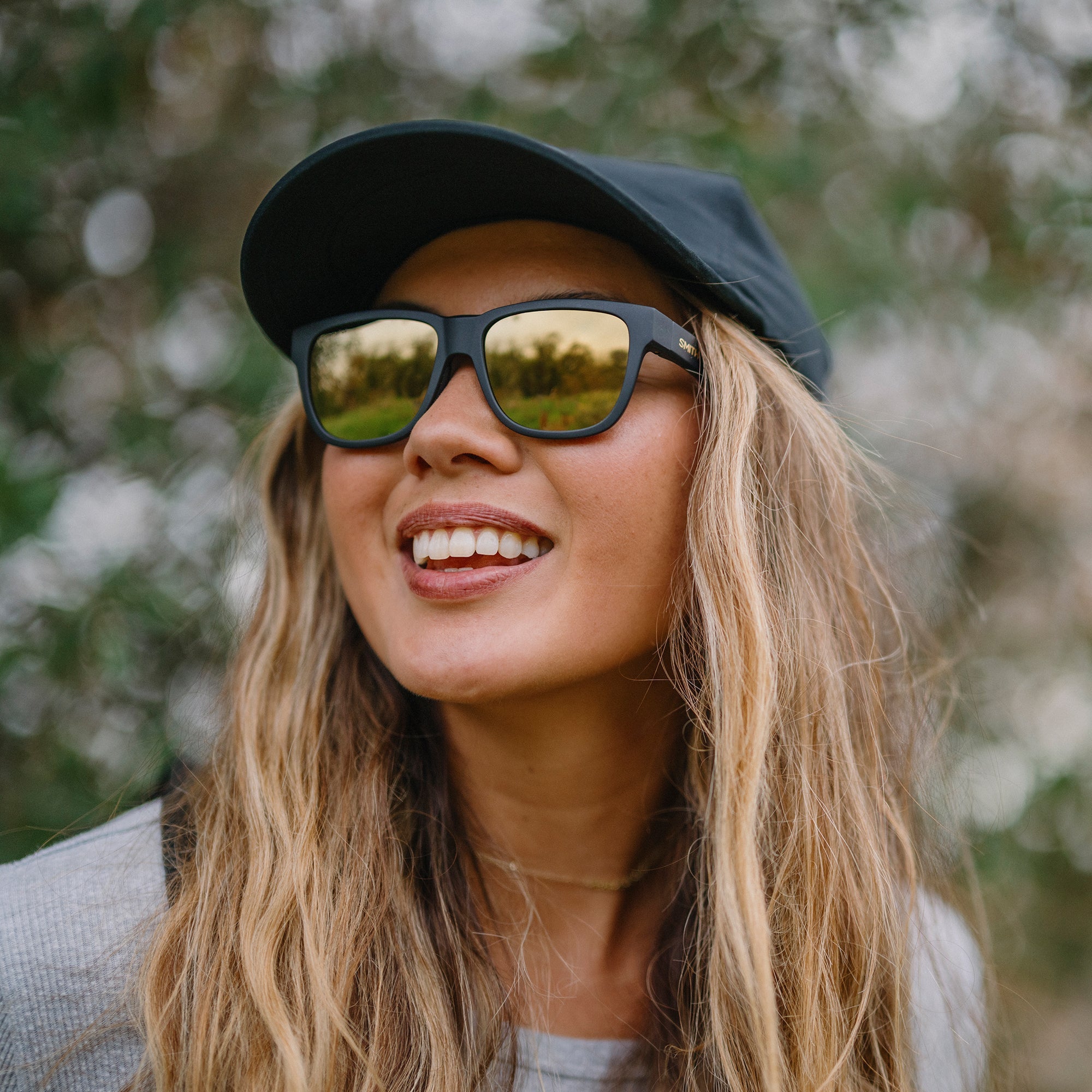Woman wearing black sunglasses with mirrored lenses and black baseball cap, smiling outdoors