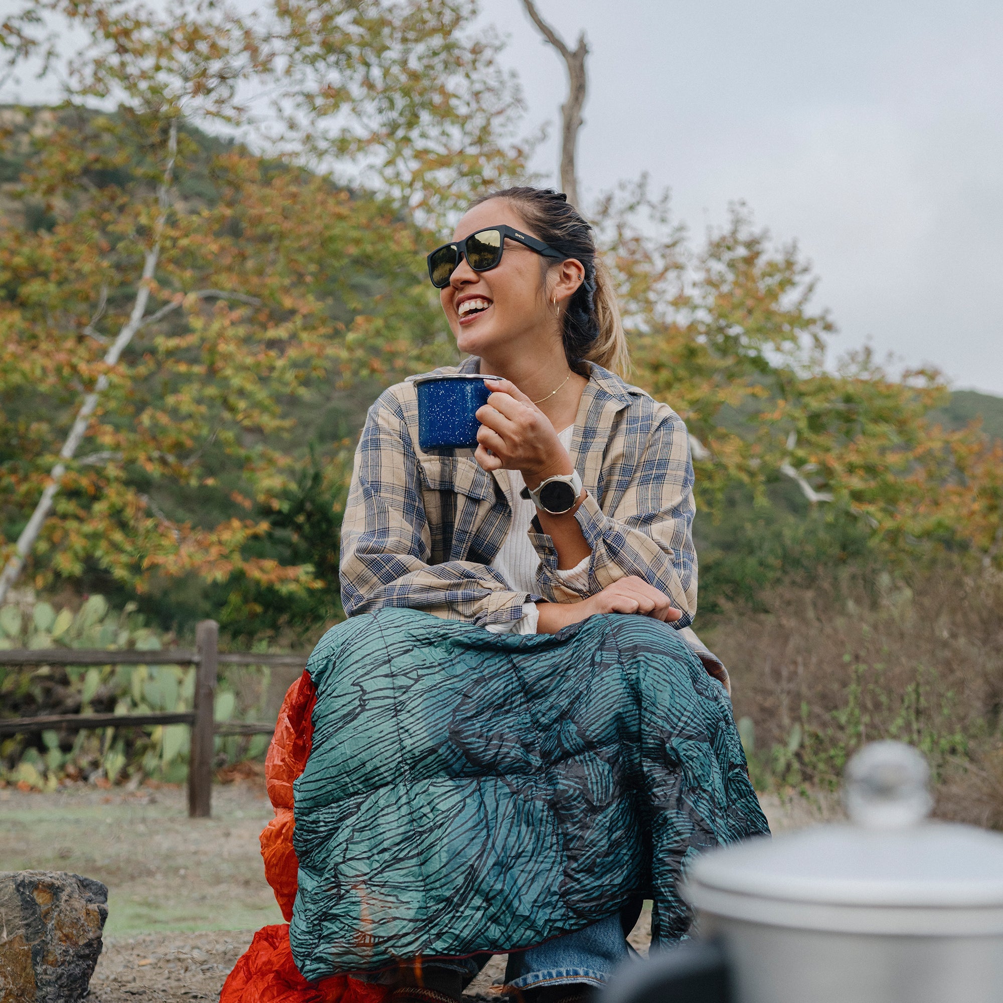 Woman wearing black sunglasses outdoors while holding blue camping mug with sleeping bag