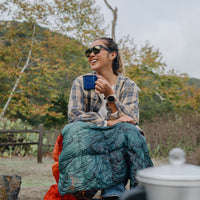 Woman wearing black sunglasses outdoors while holding blue camping mug with sleeping bag