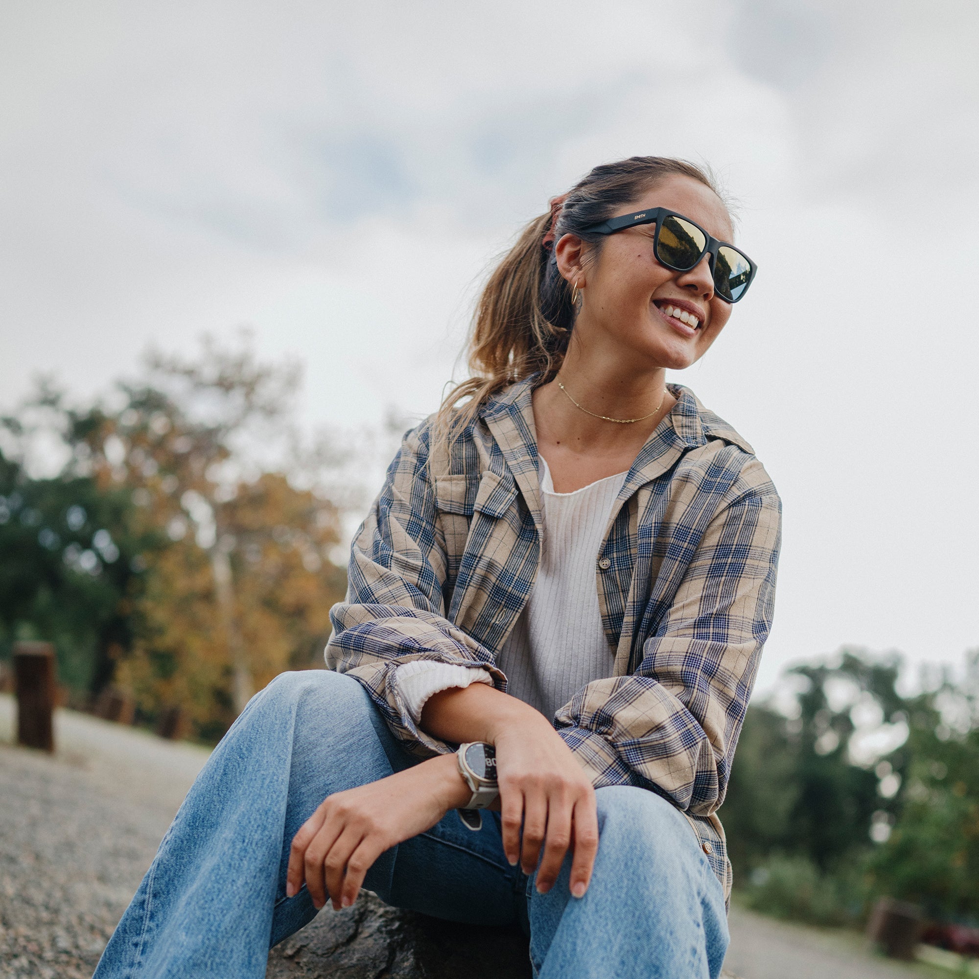 Woman wearing black polarized sunglasses with gold mirror lenses, sitting outdoors smiling
