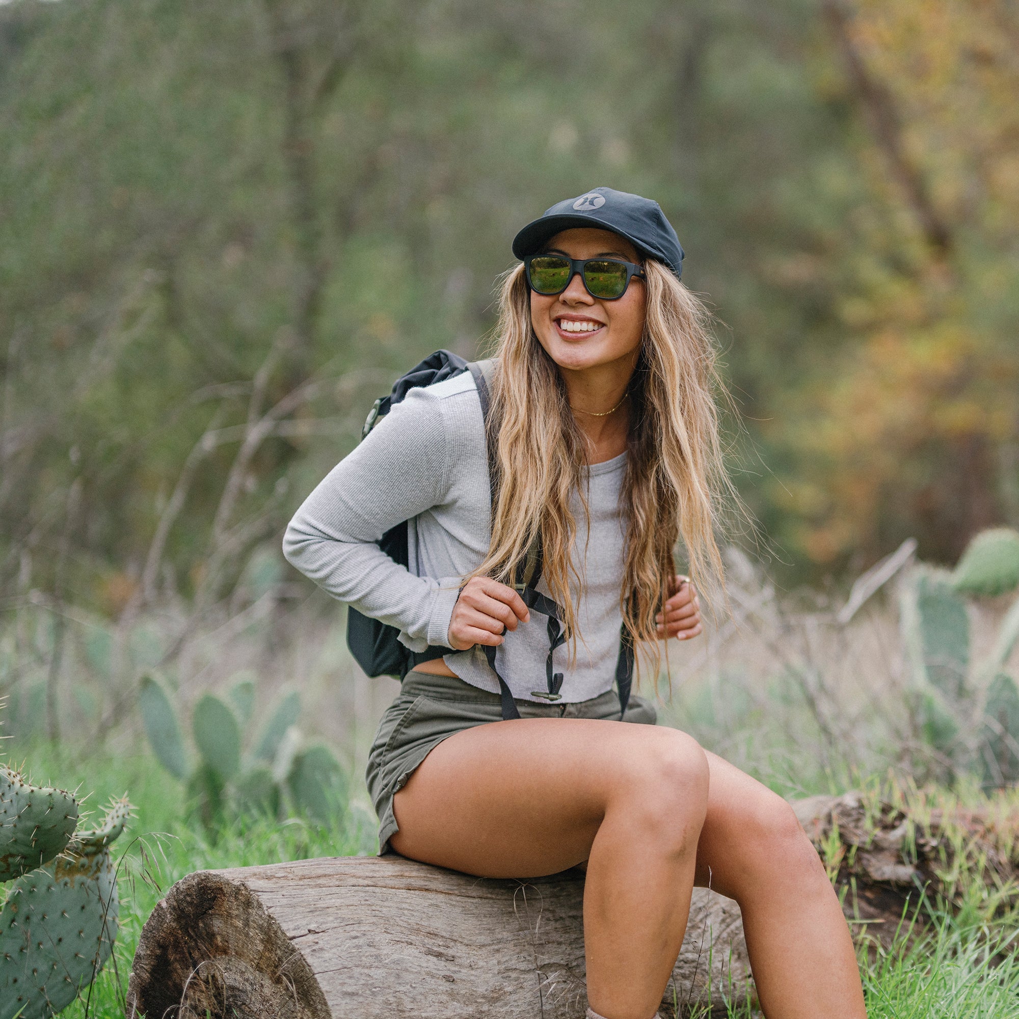 Woman wearing black sunglasses with reflective lenses sitting on log outdoors