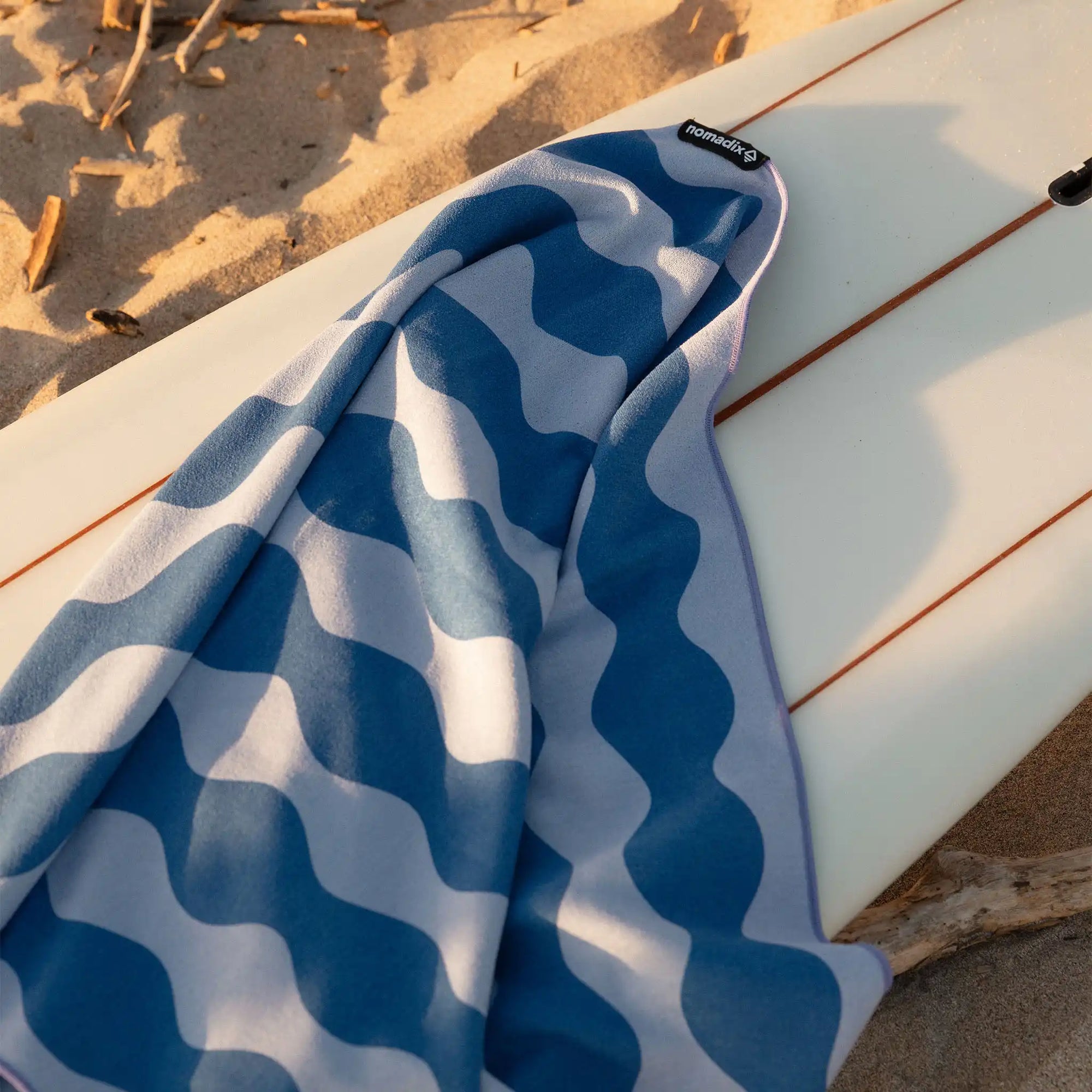 Blue and white wavy patterned towel draped over white surfboard on sandy beach