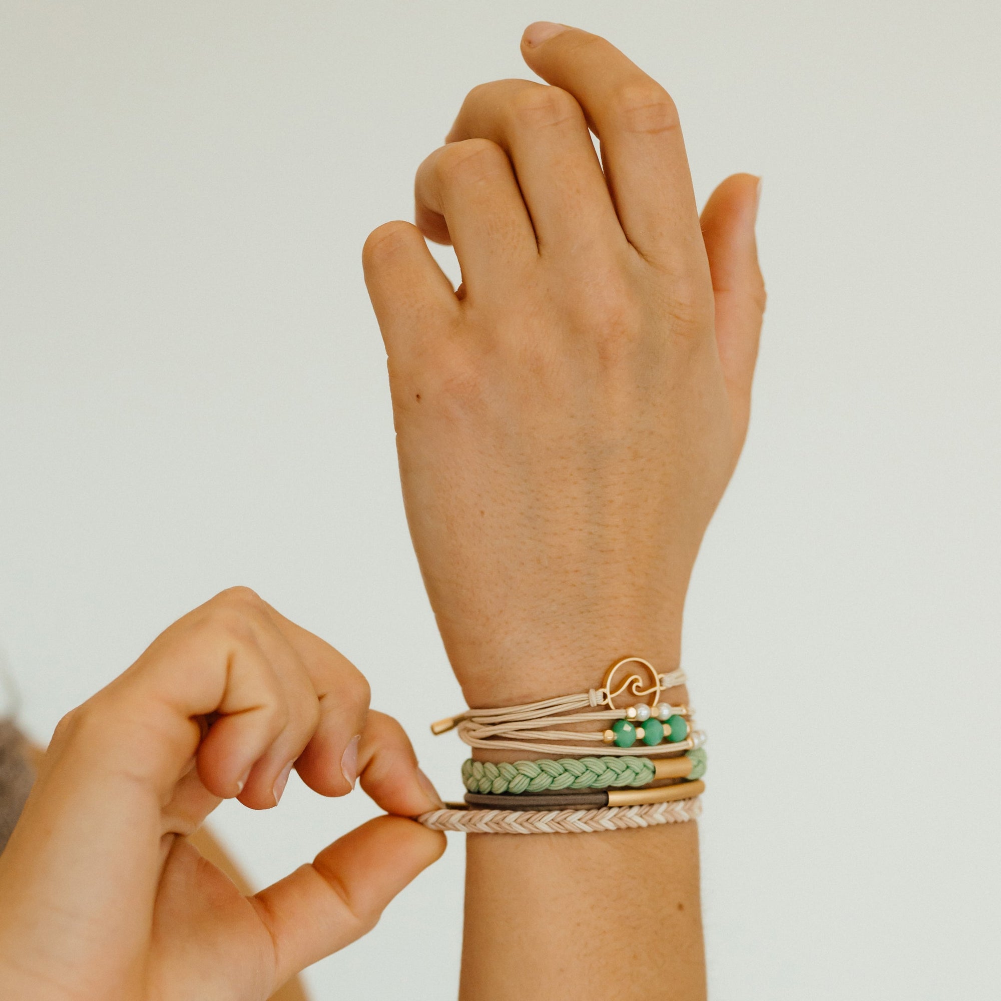 Stacked hair tie bracelets on wrist with green beads, gold wave charm, and braided cord details