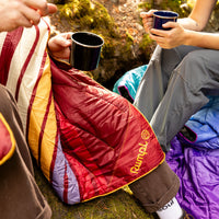 Quilted puffy blanket in retro striped pattern wrapped around camper holding metal coffee mug outdoors