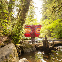 Person holding open a quilted blanket with retro concentric oval pattern in a lush forest by a stream