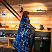 Person wrapped in navy blue paisley-print puffy blanket, back view, in rustic wood-paneled interior
