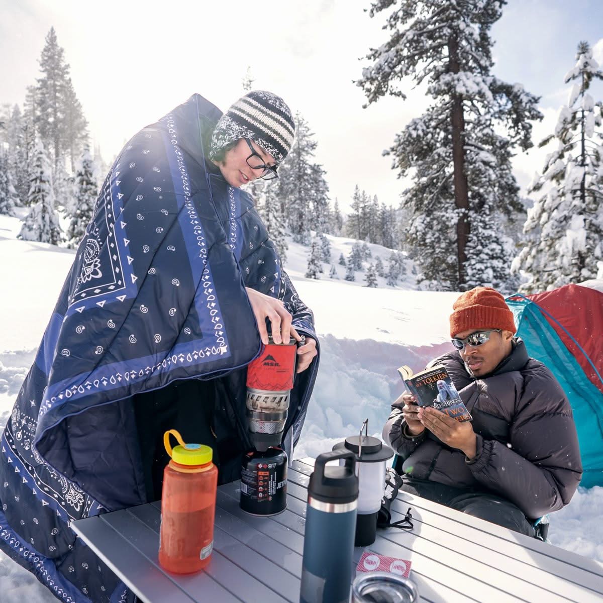 Person wrapped in navy paisley-print puffy blanket at snowy winter campsite with companion