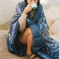 Person wrapped in navy bandana-print quilted blanket, sipping from a teal mug outdoors