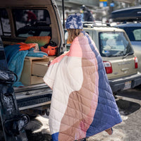 Person wrapped in gradient quilted blanket standing by open vehicle trunk outdoors
