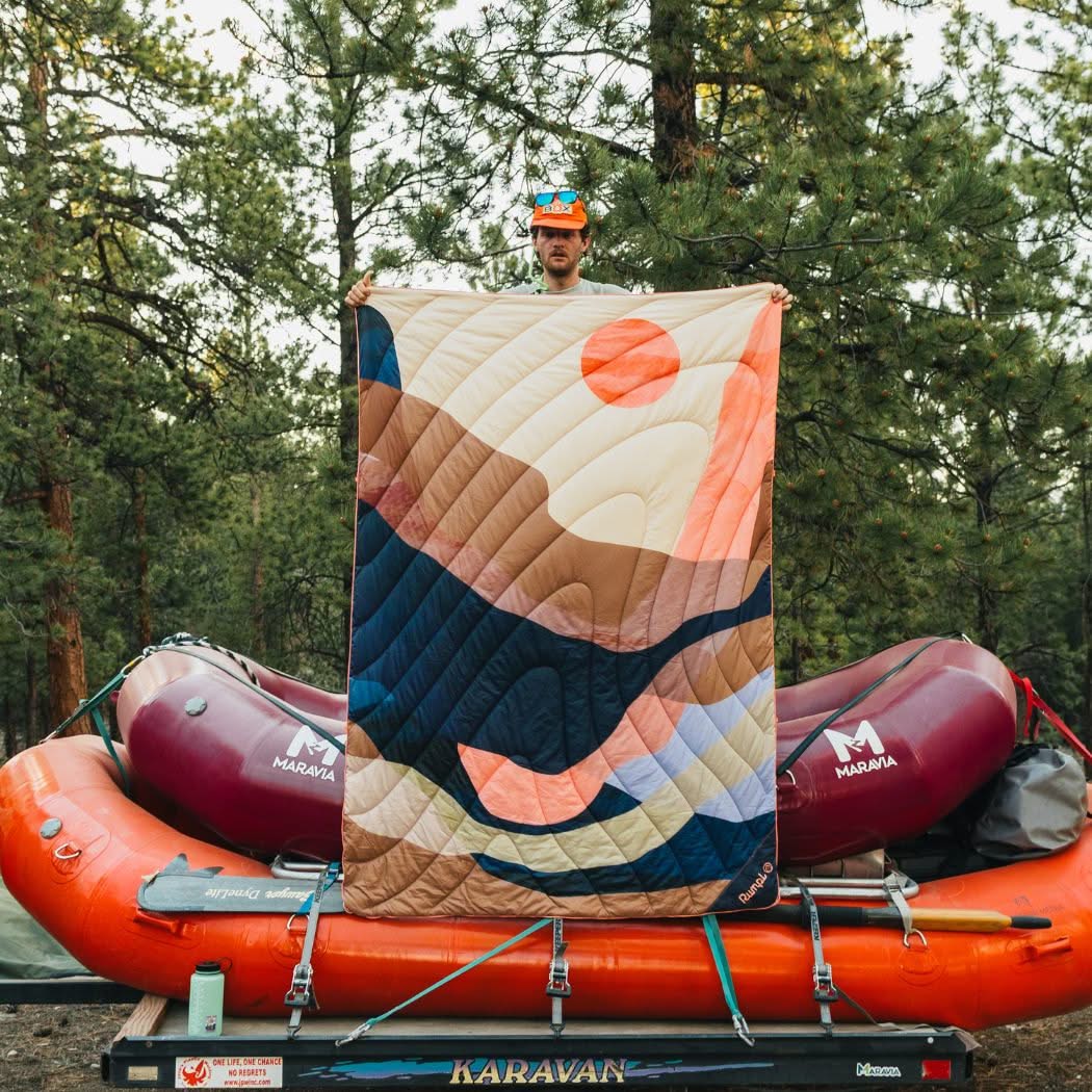 Person holding quilted outdoor blanket with sunset mountain pattern in forest camping setting