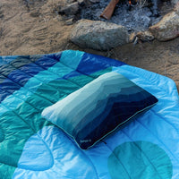 Blue gradient puffy camping blanket with matching pillow laid out near campfire