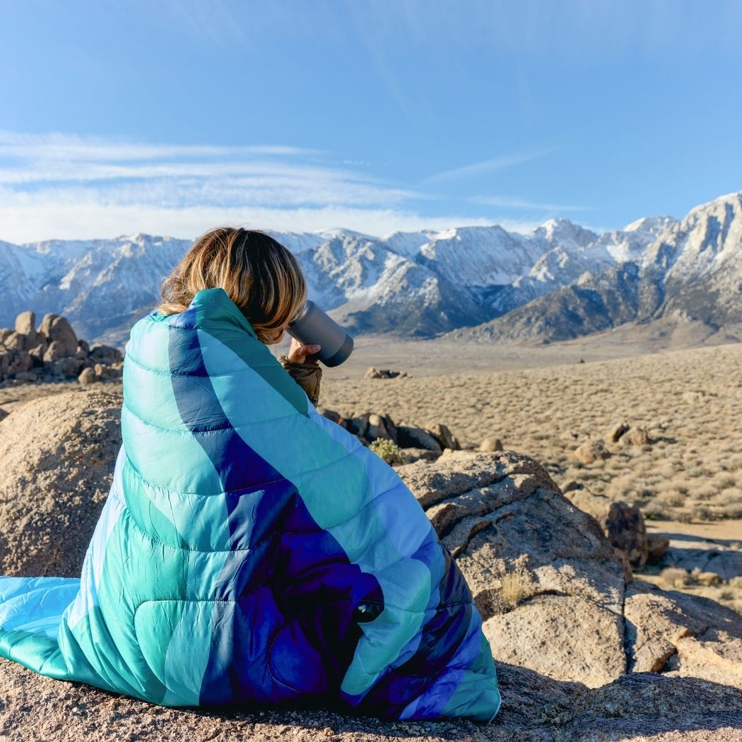 Person wrapped in blue gradient puffy blanket sitting on rocks overlooking mountain landscape