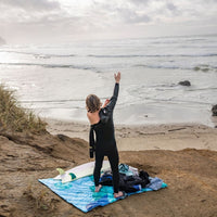 Person in wetsuit celebrating on blue patterned puffy blanket at beach with surfboard