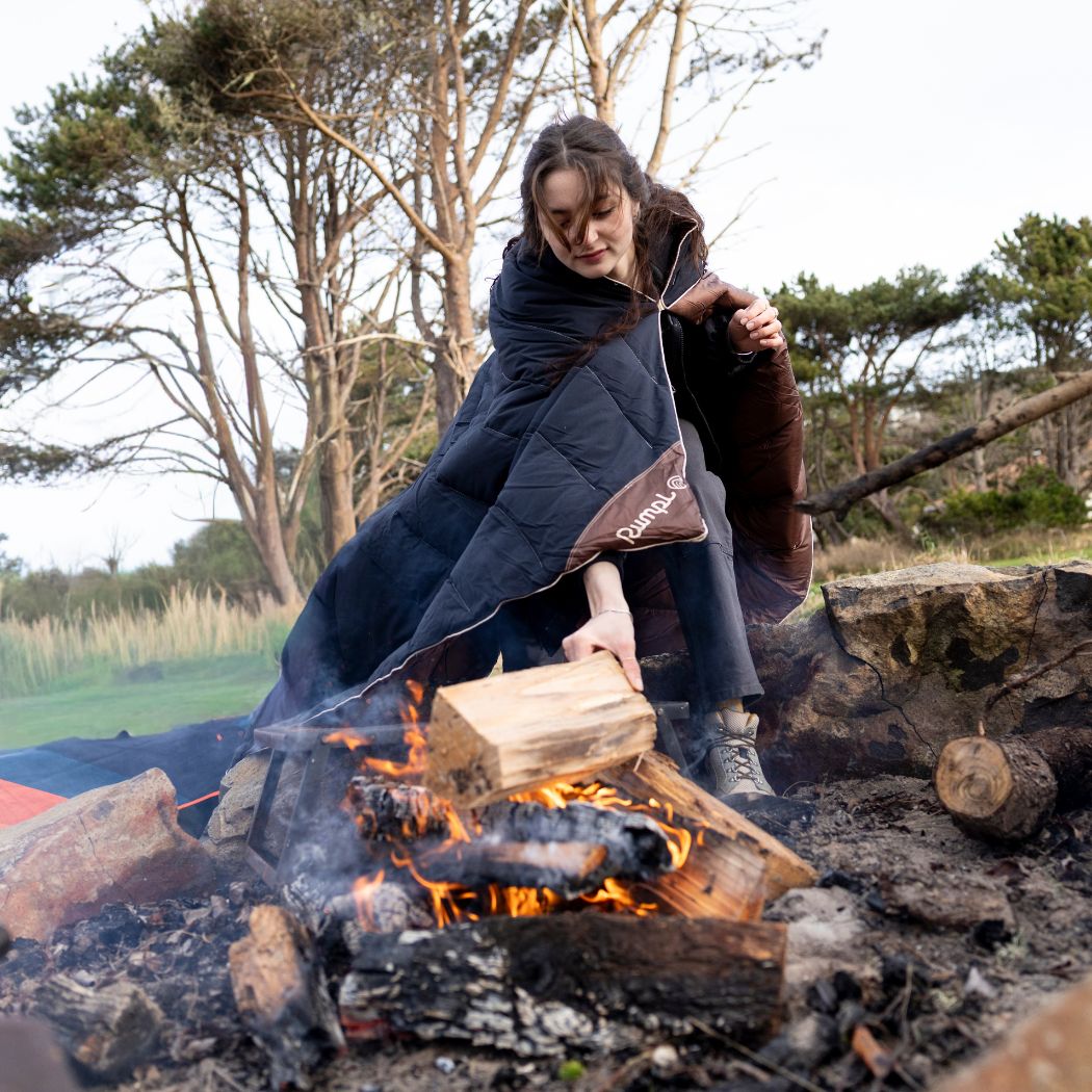Person wrapped in navy and brown puffy blanket sitting by campfire outdoors