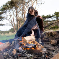 Person wrapped in navy and brown puffy blanket sitting by campfire outdoors