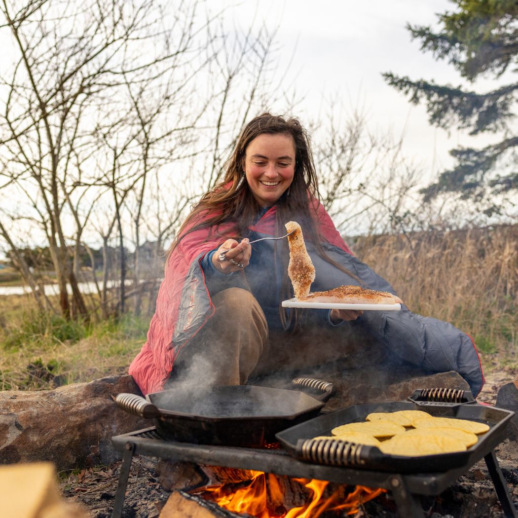 Person wrapped in coral and gray puffy blanket while cooking over campfire outdoors