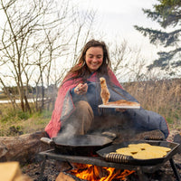 Person wrapped in coral and gray puffy blanket while cooking over campfire outdoors